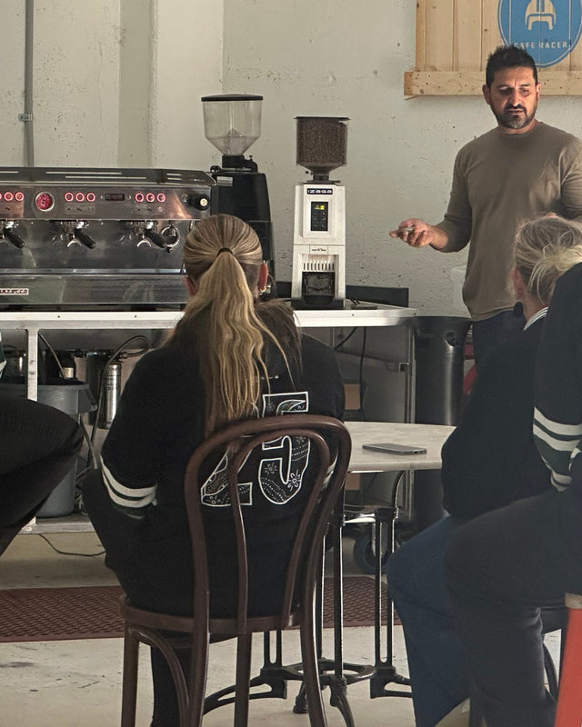 A barista teacher standing in front of students with a commercial coffee machine and grinders in the background